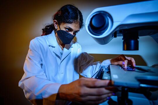 Neha Arun works with a microscope, adjusting the lens in a darkly lit lab room. She is illuminated by a blue screen. 