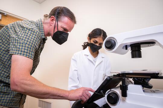 Neha Arun and Dr. Collin Kieffer, both masked, review imaging of host-pathogen interactions. The camera angle is from behind the microscope and screen, facing the two researchers head-on.